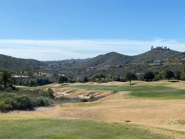 A green and flagstick beside a pond at Maderas Golf Club, with houses and hills in the distance.