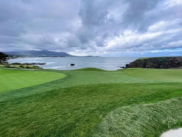 Looking down a fairway toward the ocean at Pebble Beach Golf Links, with a cypress tree on the left and a white building on the cliff.