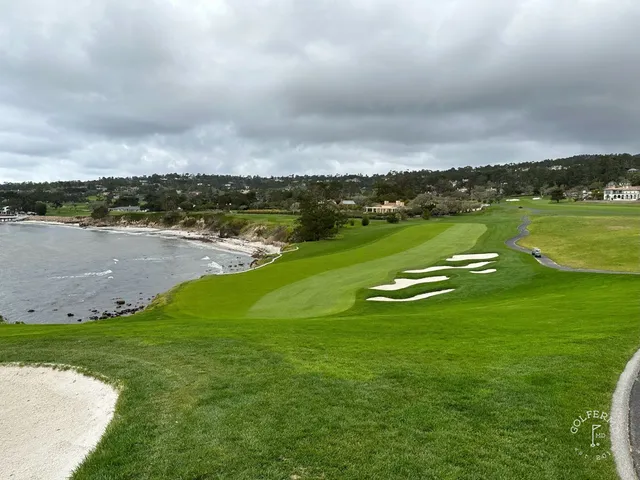 A fairway and cart path running toward the ocean at Pebble Beach Golf Links, with cypress trees and a white building on the coast.