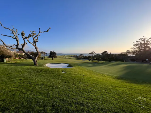 A green and flagstick beside the ocean at Pebble Beach Golf Links, with a cypress tree and a bunker.