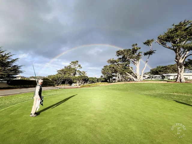 A golfer pitching toward a green beside the ocean at Pebble Beach Golf Links, with another person in the background.