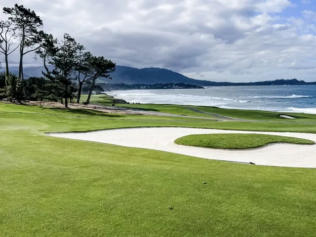 Two people walking a fairway beside the ocean at Pebble Beach Golf Links, with a cypress tree nearby.