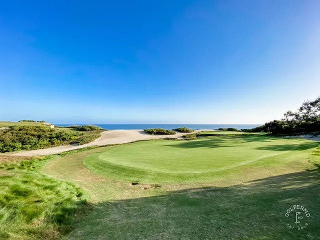 A green and flagstick beside the ocean at Pelican Hill Ocean South Course, with a cypress tree and a bunker.