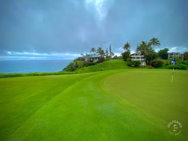A green and flagstick at Princeville Makai, seen from a rise, with the ocean and mountains beyond.
