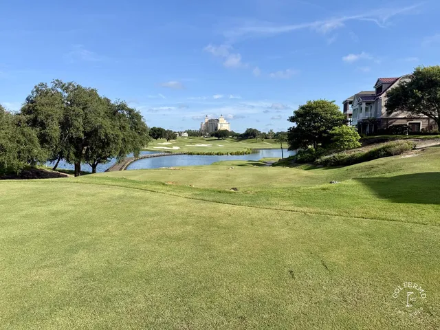 A golfer hitting a shot from the fairway at the Palmer Course, with a pond and pine trees behind the hole.