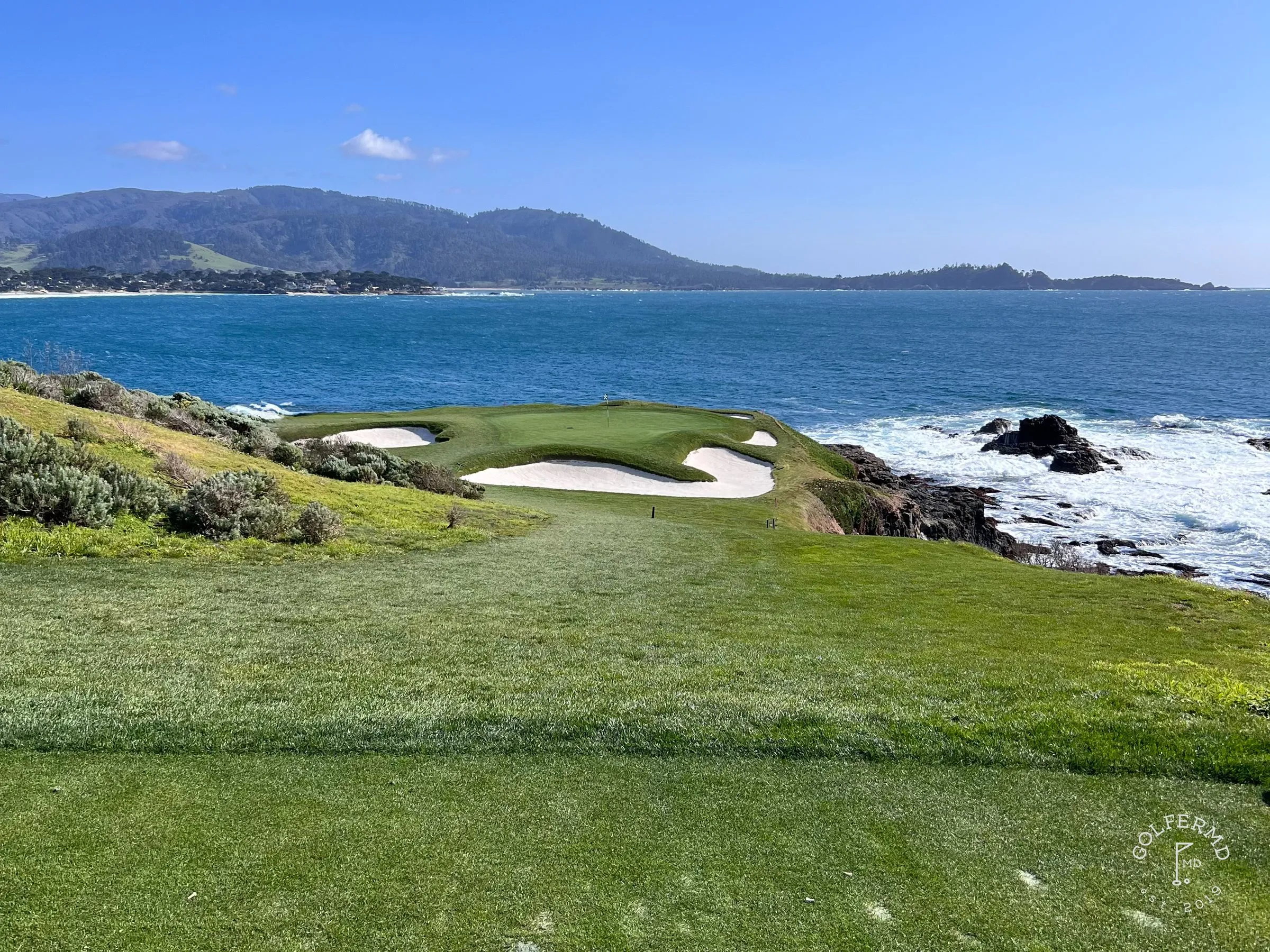 A fairway of golden grass running toward the green, with trees on the left and the ocean on the right.