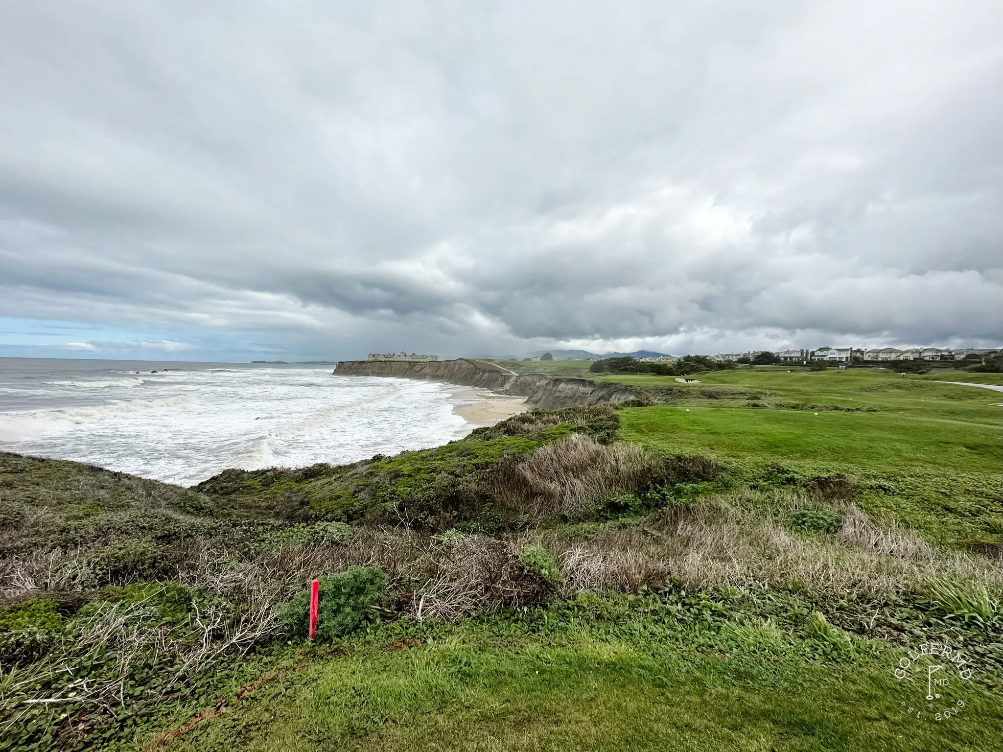 A golfer in a white cap and light shirt standing over the ball on the tee, with the ocean behind him.