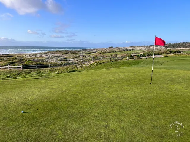 A green and flagstick in the dunes at The Links at Spanish Bay, with cypress trees and the ocean beyond.