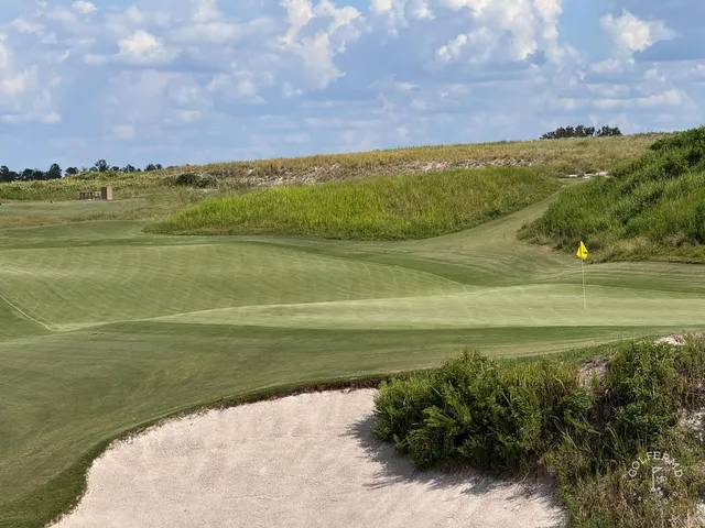 A green and flagstick at the Red Course, with a sand bunker in the foreground and dunes behind the hole.