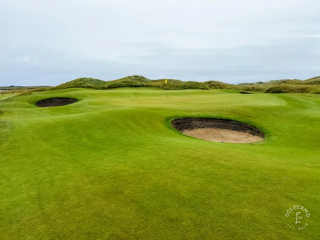 A golfer hitting an approach at Tralee, with the bay behind the green.